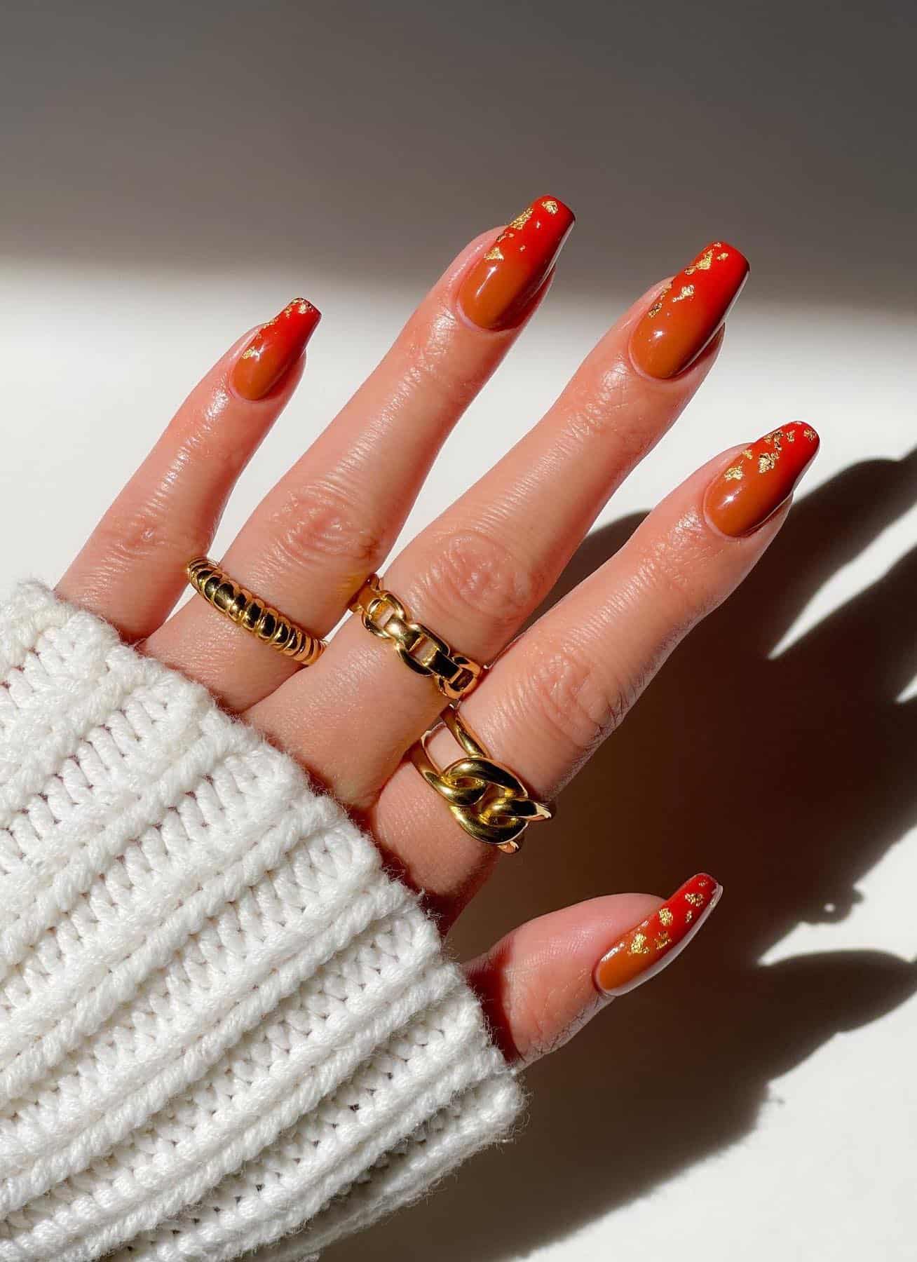 a hand with coffin nails painted a burnt orange and red ombre with gold flake accents