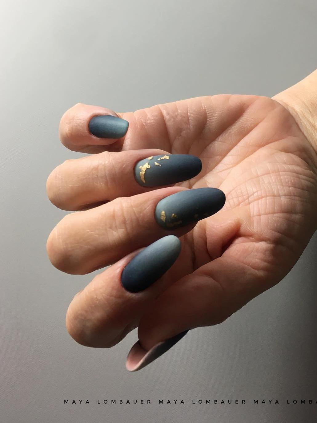 a hand with medium round nails painted light to dark green ombre with a matte finish and gold flake details