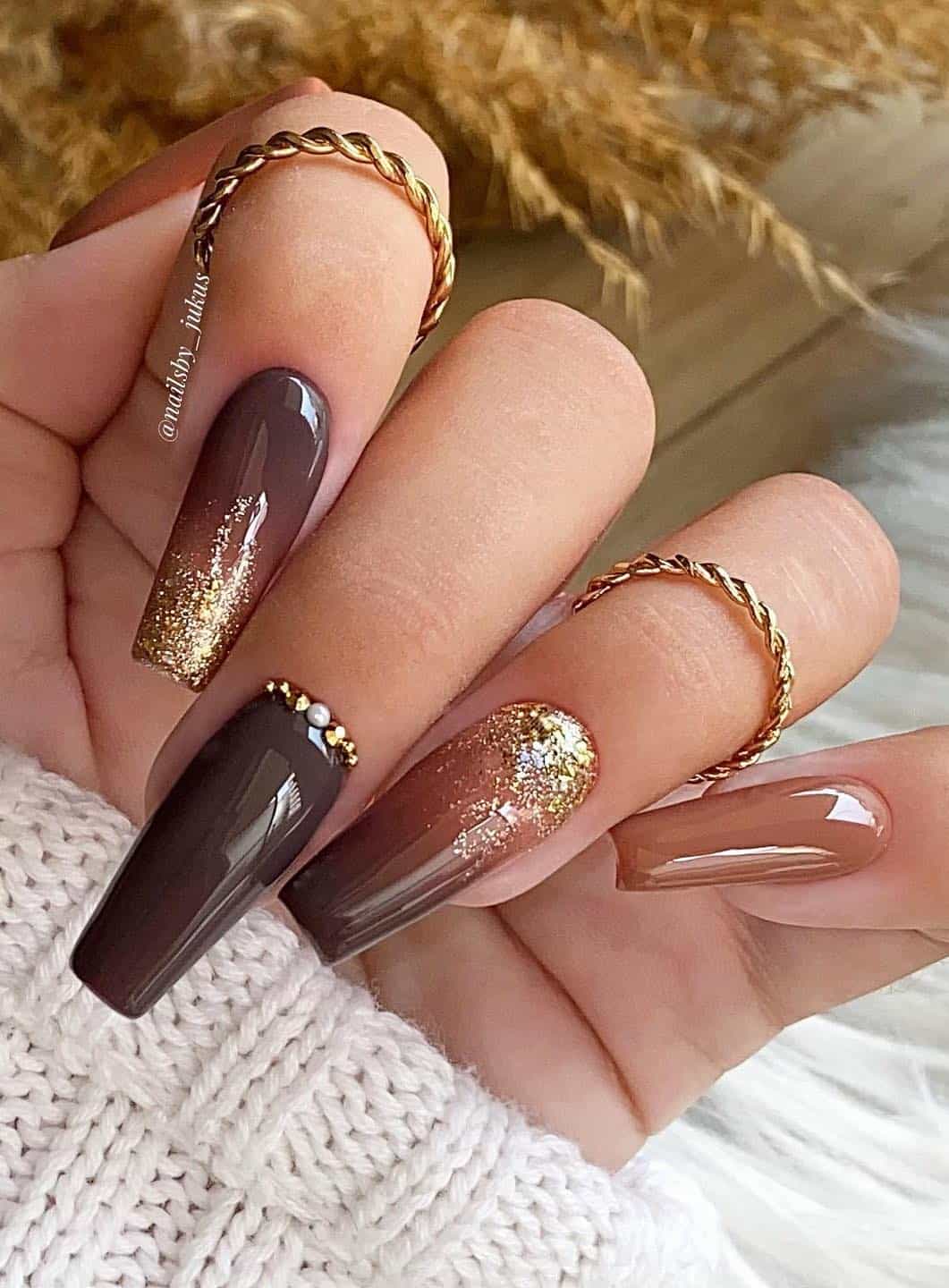 a hand with long coffin nails painted beige and brown ombre with gold glitter and gem accents
